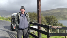 Ein Mann mit Haube und Jacke steht neben einem Zaun. Im Hintergrund ein Hügel und eine Bucht in Irland.