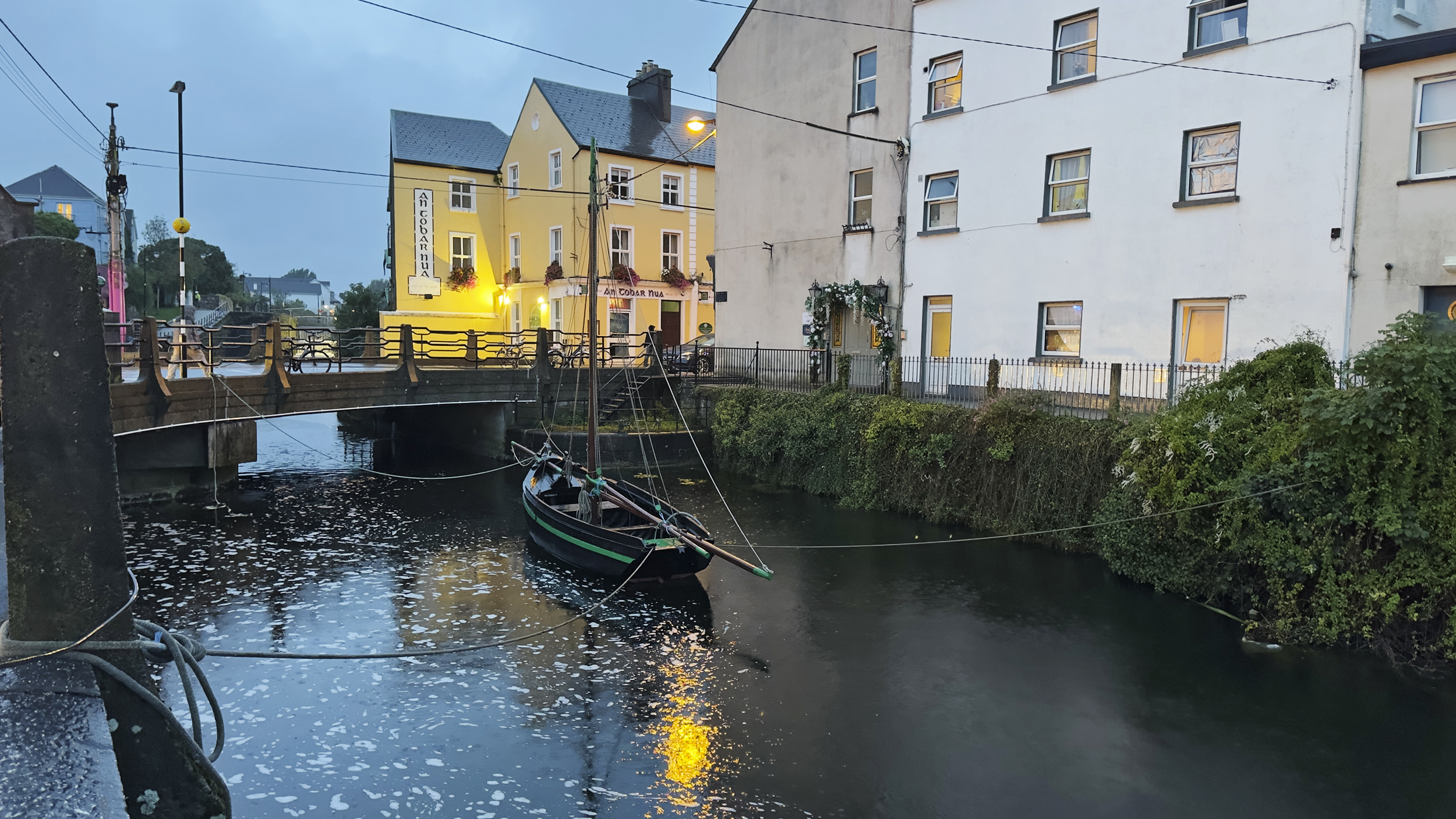 Fluss in Galway, mit kleinem Boot, Brücke und Häusern im Hintergrund.