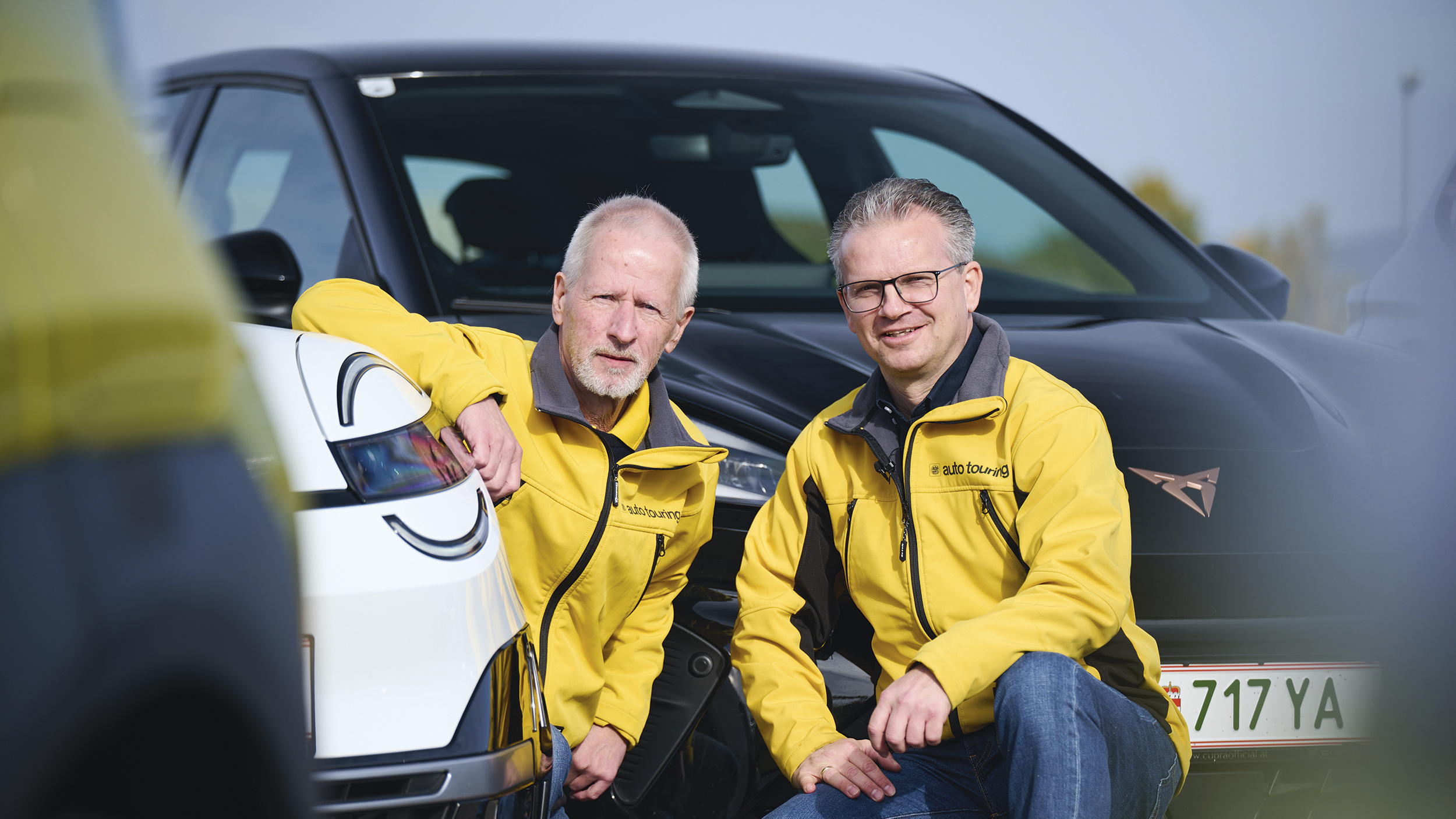 Zwei Männer in gelben autotouring-Jacken knien zwischen einem weißen Kleinwagen und einem schwarzen Cupra und blicken in die Kamera.