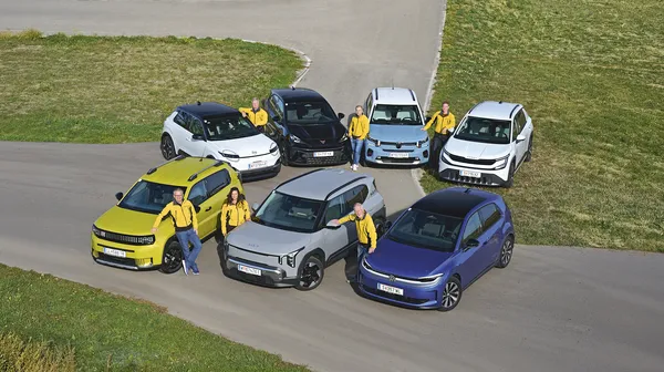 Sieben Kleinwagen in zwei Reihen auf einer asphaltierten Fläche zwischen Wiesen, aufgenommen aus erhöhter Perspektive. Sechs Personen in gelben Jacken stehen neben den Fahrzeugen.