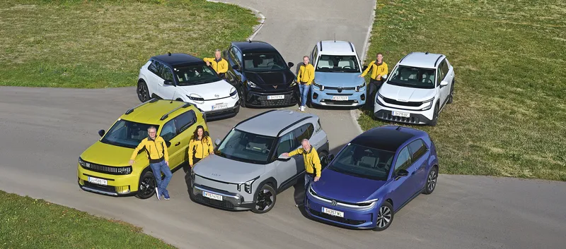 Sieben Kleinwagen in zwei Reihen auf einer asphaltierten Fläche zwischen Wiesen, aufgenommen aus erhöhter Perspektive. Sechs Personen in gelben Jacken stehen neben den Fahrzeugen.