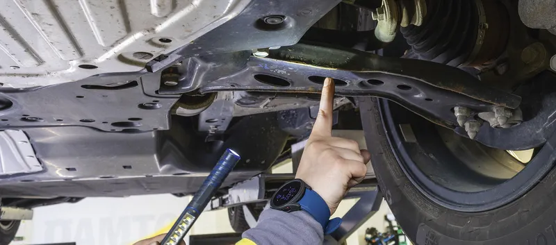 Jemand prüft den Unterboden eines Autos auf einer Hebebühne und zeigt mit dem Finger auf einen Querlenker der Vorderradaufhängung. In der anderen Hand hält er eine Inspektionsleuchte.