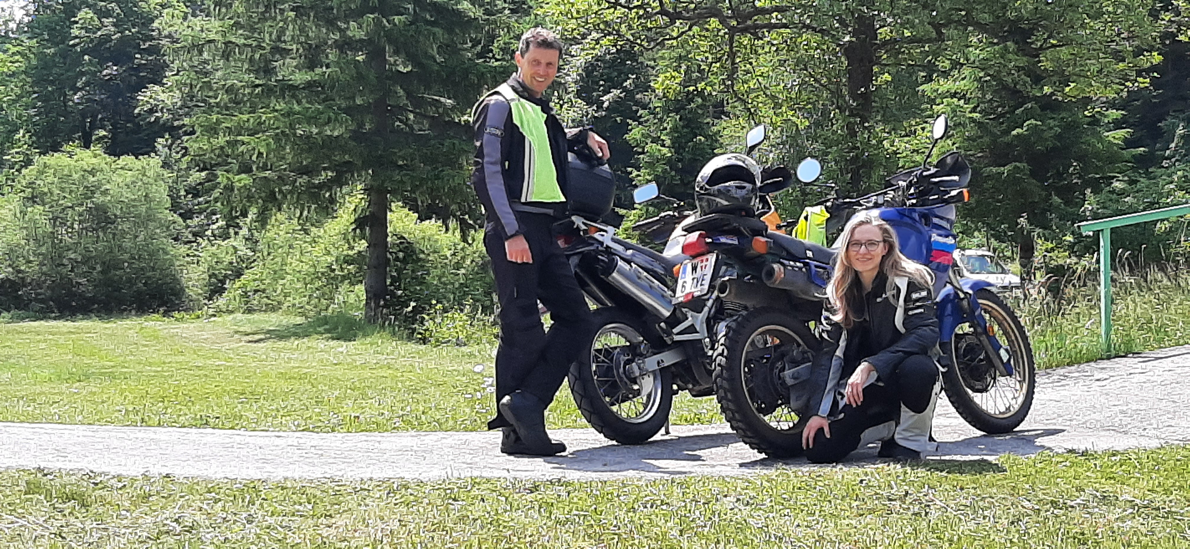 Auf dem Bild sieht man zwei Personen mit ihren Motorrädern. Ein Mann steht neben seinem Motorrad. Eine Frau kniet vor ihrem Bike.