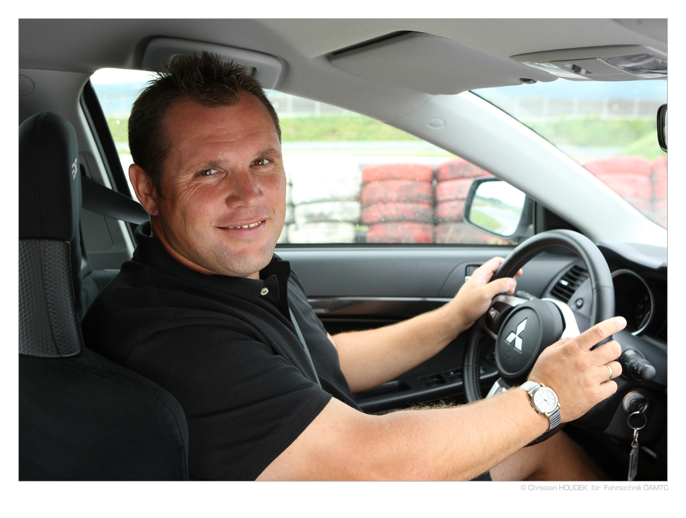 Das Foto zeigt einen Mann der hinter dem Steuer eines Autos sitzt und freundlich in die Kamera lächelt.