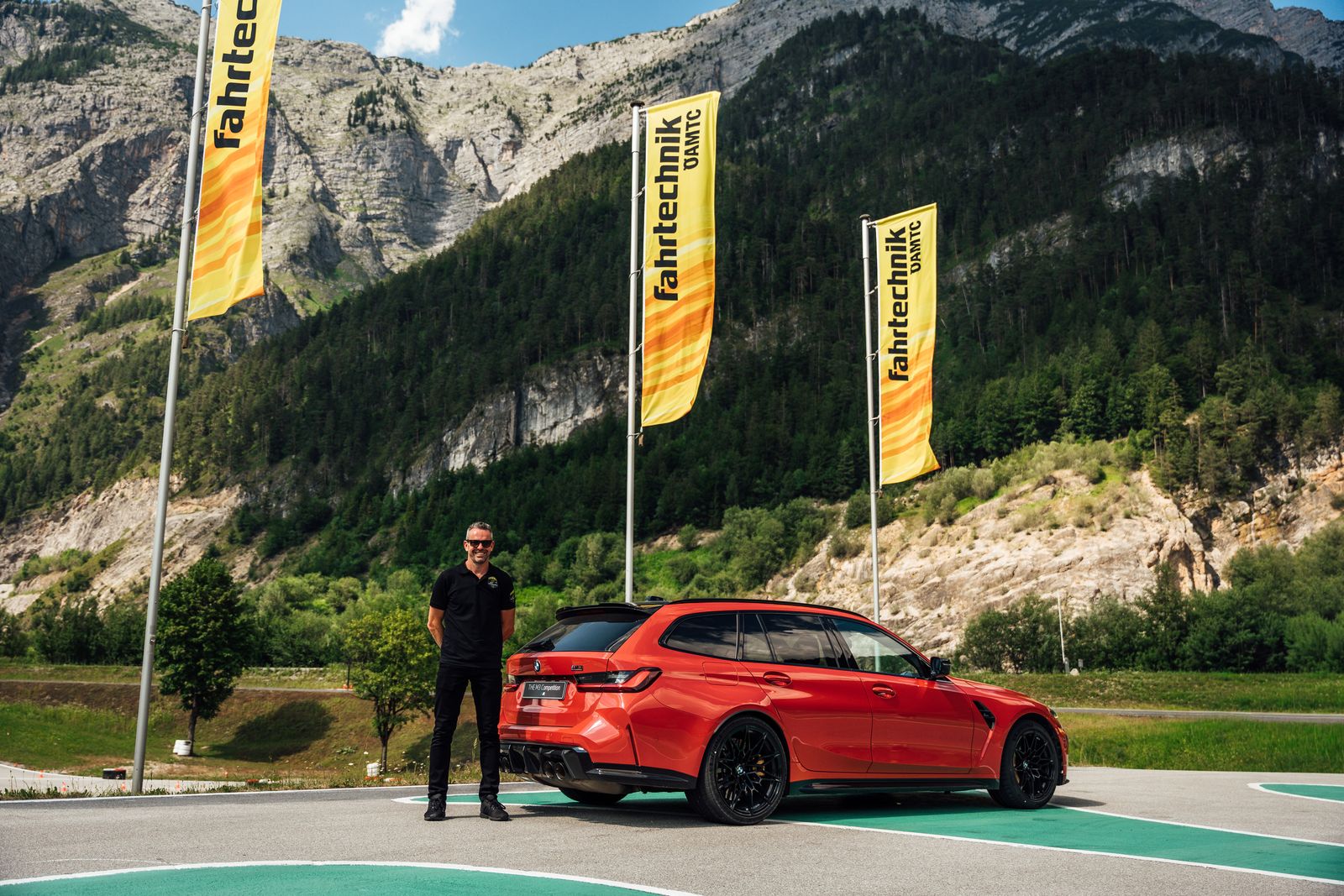 Es ist ein Mann zu sehen, der neben einem roten Auto steht. Im Hintergrund sind gelbe Fahnen und Berge zu sehen.