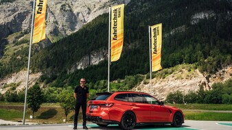Es ist ein Mann zu sehen, der neben einem roten Auto steht. Im Hintergrund sind gelbe Fahnen und Berge zu sehen.