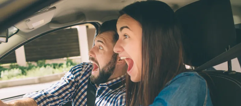 Es ist ein Mann und eine Frau zu sehen, die im Auto sitzen und erschrocken schauen.