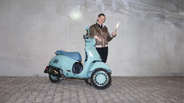 Mann in brauner Jacke steht neben einer türkisfarbenen Vespa vor einer Betonwand und hält eine sprühende Wunderkerze.
