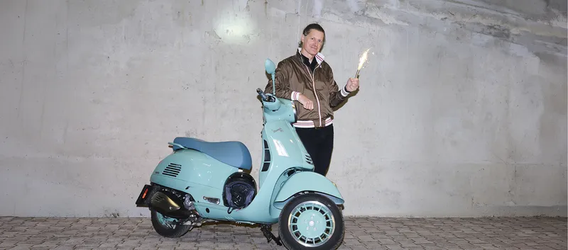 Mann in brauner Jacke steht neben einer türkisfarbenen Vespa vor einer Betonwand und hält eine sprühende Wunderkerze.
