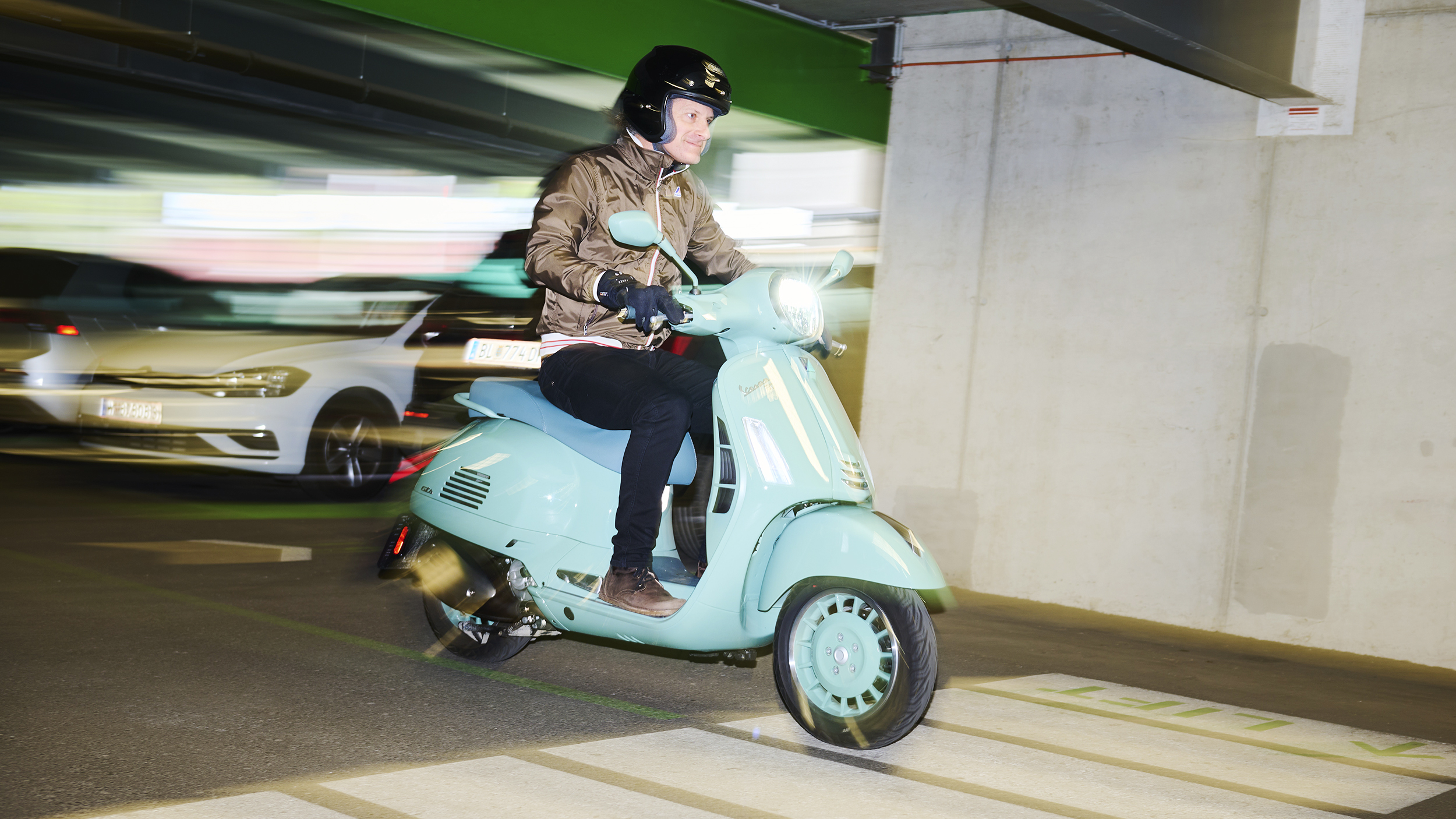Mann mit schwarzem Helm, Handschuhen und brauner Jacke fährt eine türkisfarbene Vespa mit eingeschaltetem Scheinwerfer durch eine Tiefgarage.