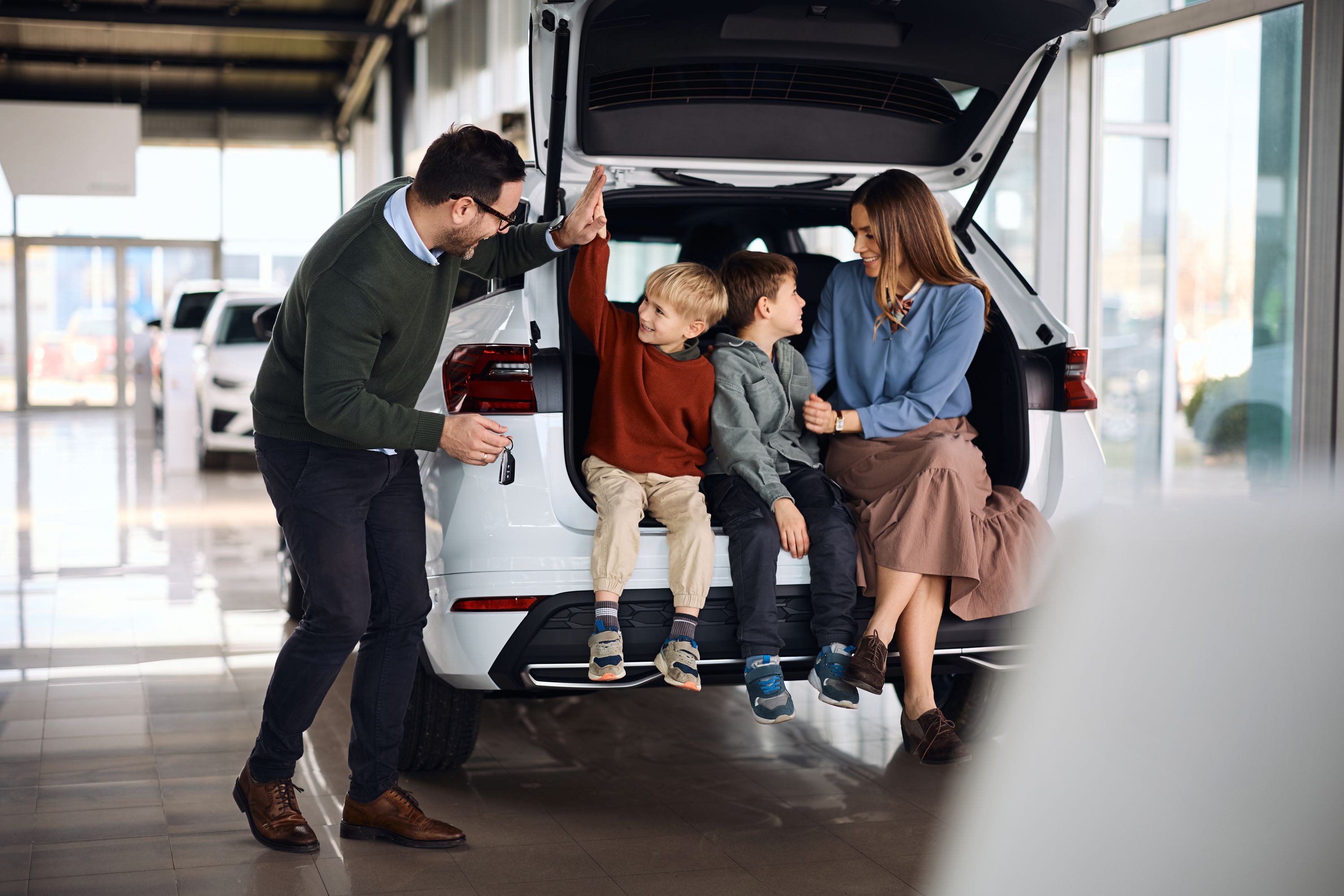 Eine fröhliche Familie mit zwei Söhnen sitzt im Kofferraum von einem weißen Neuwagen.