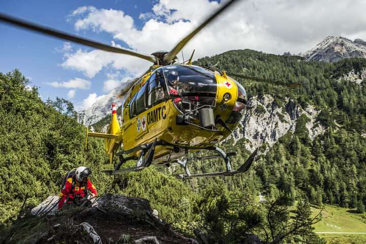 Ein ÖAMTC-Rettungshubschrauber fliegt in einer gebirgigen Umgebung sehr nah am Boden, während ein Sanitäter am Boden steht.