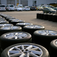 Ein Schmuckbild zum ÖAMTC-Sommerreifentest 2026: Zwei Reihen übereinander gestapelter Autoreifen vor einem Gebäude, vor dem Autos geparkt sind.