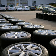 Ein Schmuckbild zum ÖAMTC-Sommerreifentest 2026: Zwei Reihen übereinander gestapelter Autoreifen vor einem Gebäude, vor dem Autos geparkt sind.