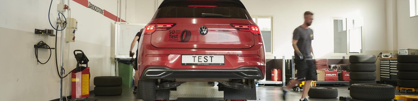 Sommerreifentest - Auto in Werkstatt ist auf der Hebebühne, rechts im Bild sieht man einzelne Reifen, Werkzeug und einen Mann.