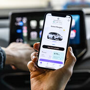 Das Foto zeigt eine Hand, die ein Smartphone hält. Auf dem Display des Smartphones sieht man ein Auto. Dahinter sieht man das Display am Armaturenbrett eines Autos.
