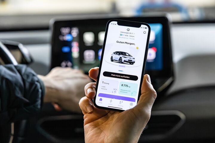 Das Foto zeigt eine Hand, die ein Smartphone hält. Auf dem Display des Smartphones sieht man ein Auto. Dahinter sieht man das Display am Armaturenbrett eines Autos.