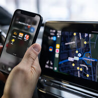Das Foto zeigt eine Hand, die ein Smartphone hält. Auf dem Display des Smartphones sind verschiedene Apps zu erkennen. Rechts vom Smartphone sieht man das Display am Amaturenbrett eines Autos. Darauf ist eine Straßenkarte zu sehen.