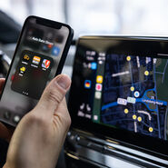 Das Foto zeigt eine Hand, die ein Smartphone hält. Auf dem Display des Smartphones sind verschiedene Apps zu erkennen. Rechts vom Smartphone sieht man das Display am Amaturenbrett eines Autos. Darauf ist eine Straßenkarte zu sehen.