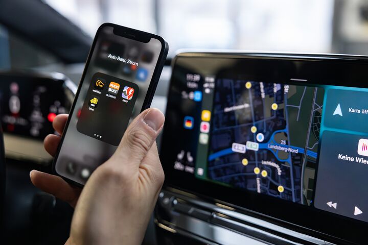 Das Foto zeigt eine Hand, die ein Smartphone hält. Auf dem Display des Smartphones sind verschiedene Apps zu erkennen. Rechts vom Smartphone sieht man das Display am Amaturenbrett eines Autos. Darauf ist eine Straßenkarte zu sehen.