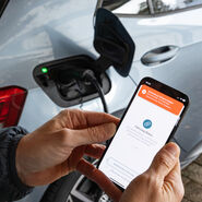 Das Foto zeigt zwei Hände, die ein Smartphone halten. Auf dem Display des Smartphones sieht man den Text "Fahrzeug Status". Links daneben sieht man den geöffneten Tankdeckel eines Elektroautos, das gerade mit einem Stromkabel geladen wird.