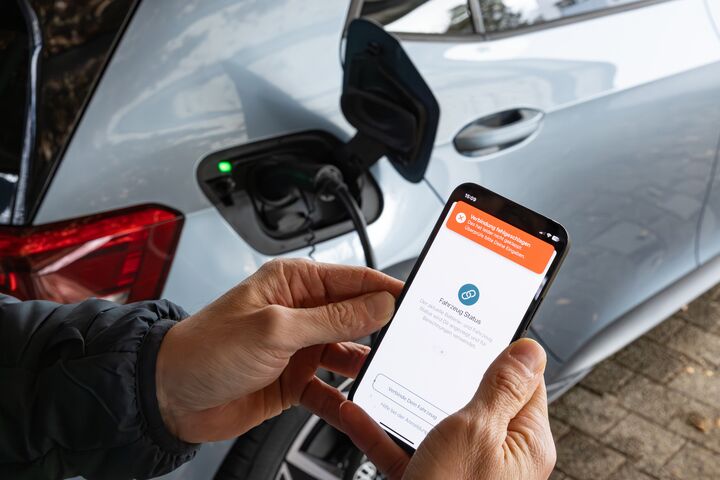 Das Foto zeigt zwei Hände, die ein Smartphone halten. Auf dem Display des Smartphones sieht man den Text "Fahrzeug Status". Links daneben sieht man den geöffneten Tankdeckel eines Elektroautos, das gerade mit einem Stromkabel geladen wird.