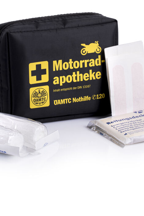 ÖAMTC Motorradapotheke in schwarzer Weichverpackung mit gelber Schrift und davor liegen Verbände, eine Rettungsdecke und Pflasterstrips.