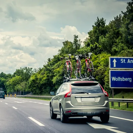Auf einer Autobahn fährt ein graues Auto mit drei Fahrrädern am Dachträger. Neben der Straße sind grüne Bäume und der Himmel ist bewölkt.