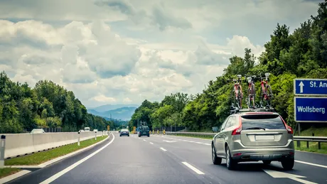 Auf einer Autobahn fährt ein graues Auto mit drei Fahrrädern am Dachträger. Neben der Straße sind grüne Bäume und der Himmel ist bewölkt.