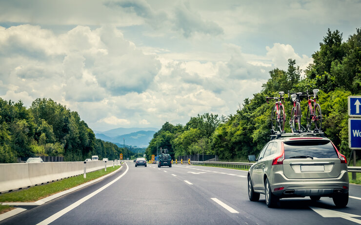 Auf einer Autobahn fährt ein graues Auto mit drei Fahrrädern am Dachträger. Neben der Straße sind grüne Bäume und der Himmel ist bewölkt.