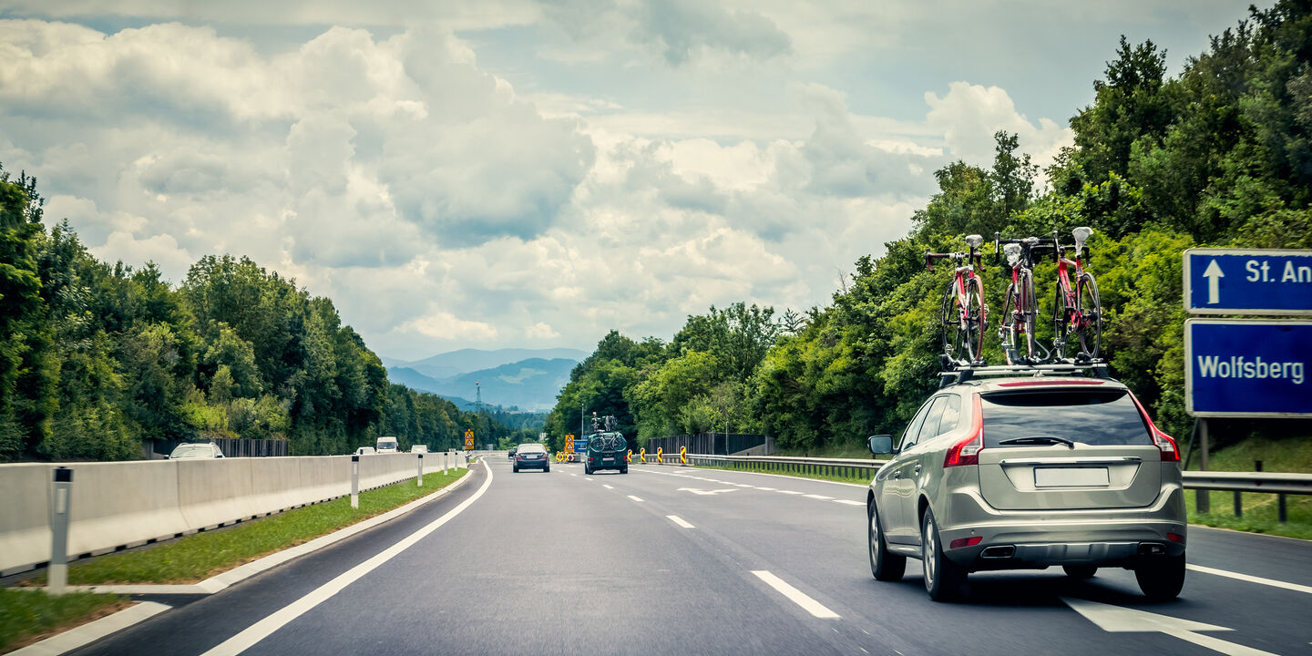 Auf einer Autobahn fährt ein graues Auto mit drei Fahrrädern am Dachträger. Neben der Straße sind grüne Bäume und der Himmel ist bewölkt.