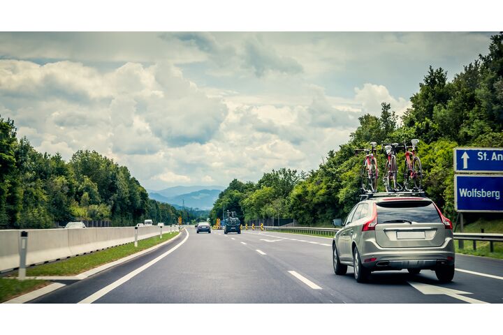 Auf einer Autobahn fährt ein graues Auto mit drei Fahrrädern am Dachträger. Neben der Straße sind grüne Bäume und der Himmel ist bewölkt.