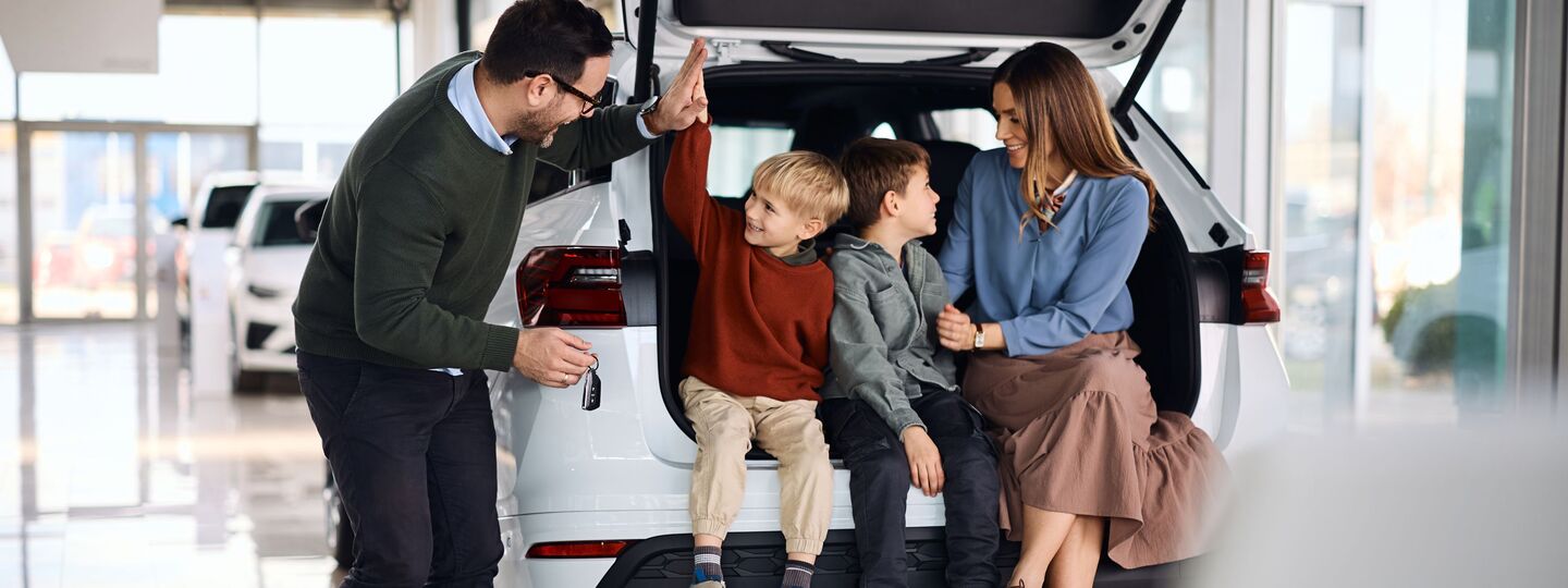Eine fröhliche Familie mit zwei Söhnen sitzt im Kofferraum von einem weißen Neuwagen.