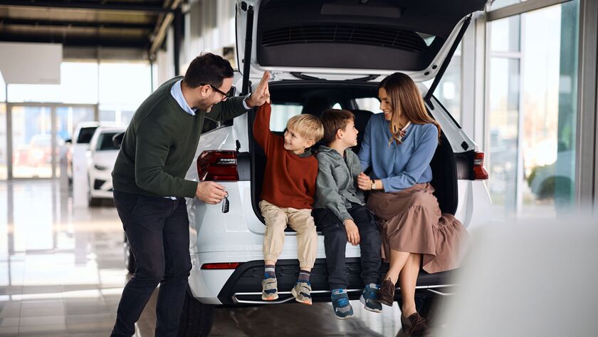 Eine fröhliche Familie mit zwei Söhnen sitzt im Kofferraum von einem weißen Neuwagen.