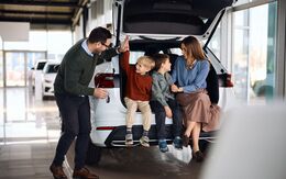 Eine fröhliche Familie mit zwei Söhnen sitzt im Kofferraum von einem weißen Neuwagen.