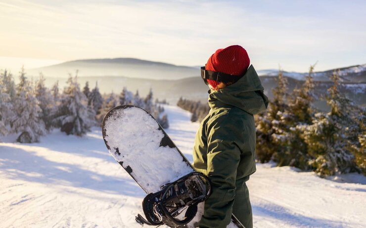 Snowboarderin steht auf Skipiste mit verschneitem Snowboard in der Hand, nur mit Mütze und ohne Skihelm.