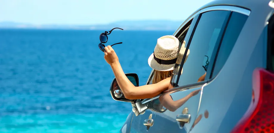 Eine Frau mit Strohhut hält ihre Sonnenbrille aus dem Autofenster und blickt aufs Meer.