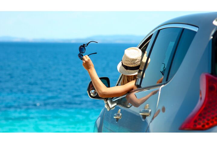 Eine Frau mit Strohhut hält ihre Sonnenbrille aus dem Autofenster und blickt aufs Meer.