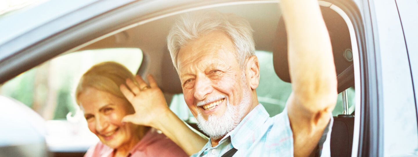 Ein älteres Paar in Großaufnahme sitzt im Auto winkt zufrieden hinaus. 