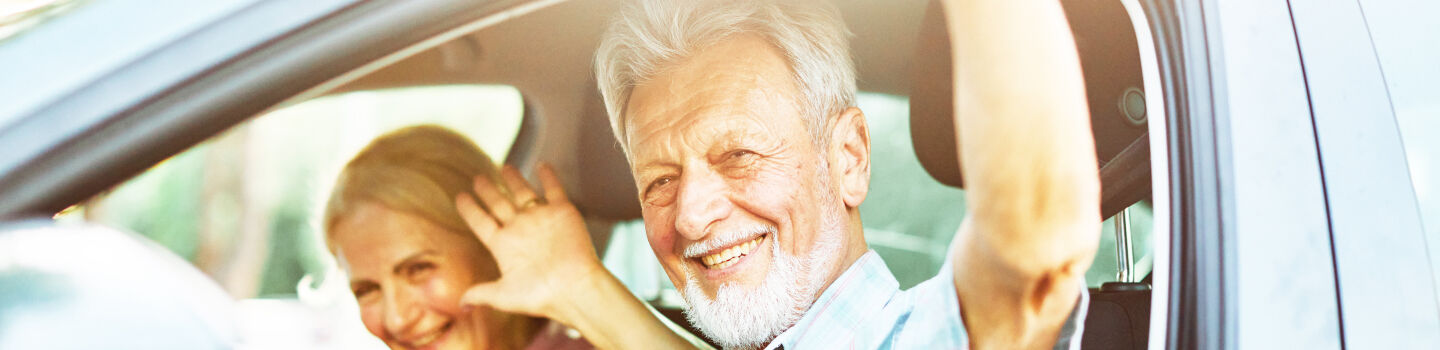 Ein älteres Paar in Großaufnahme sitzt im Auto winkt zufrieden hinaus. 
