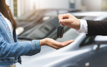 Eine Hand hält einen Autoschlüssel und gibt ihn einer anderen Person weiter. Im Hintergrund stehen zwei Autos.