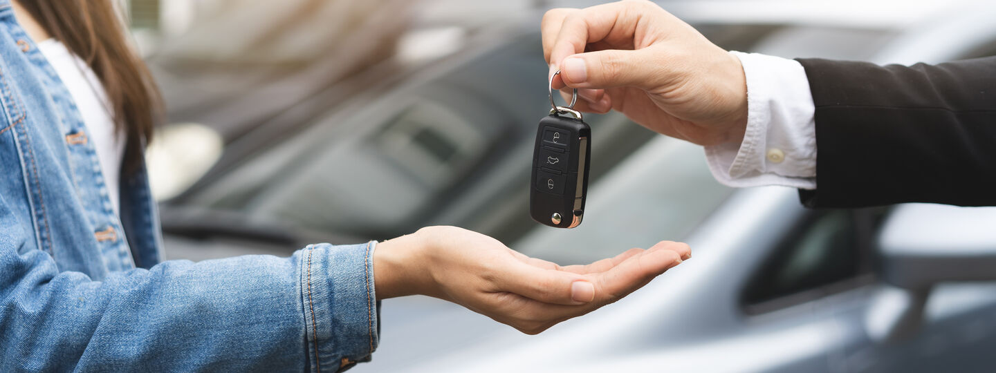 Eine Hand hält einen Autoschlüssel und gibt ihn einer anderen Person weiter. Im Hintergrund stehen zwei Autos.