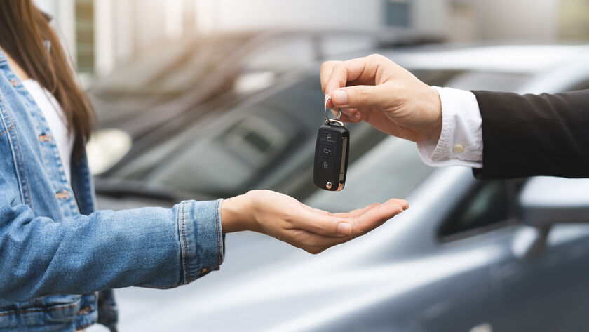 Eine Hand hält einen Autoschlüssel und gibt ihn einer anderen Person weiter. Im Hintergrund stehen zwei Autos.
