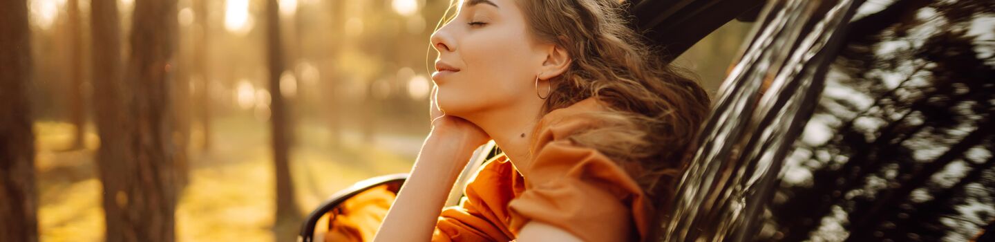Ein dunkles Auto steht im Wald, die Sonne scheint durch den herbstlichen Wald. Eine Frau sitzt entspannt am Fahrersitz und lehnt sich gemütlich aus dem geöffneten Fenster, sie hat die Augen geschlossen und genießt die Situation.