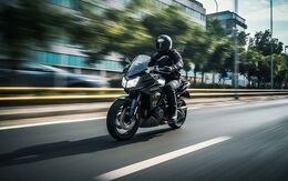 Ein Motorradfahrer in der Stadt, mit Helm und vollständiger Ausrüstung, fährt auf einer Straße. Im Hintergrund ein großes Bürogebäude und Bäume.