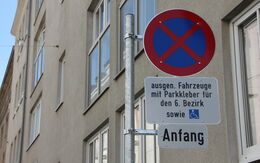 Das Bild zeigt ein Parkverbotsschild mit zwei Zusatztafeln in Wien. Auf dem oberen Zusatzschild steht: "ausgen. Fahrzeuge mit Parkkleber für den 6. Bezirk sowie" und zeigt ein Symbol für einen Behindertenparkplatz. Das untere Zusatzschild zeigt das Wort "Anfang“.