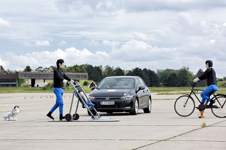 Ein schwarzes Auto am Testgelände, vor dem Fahrzeug stellen Dummies Fußgänger mit Kinderwagen und einen Fahrradfahrer dar. Ein Hundedummie ist auch vor dem Auto.