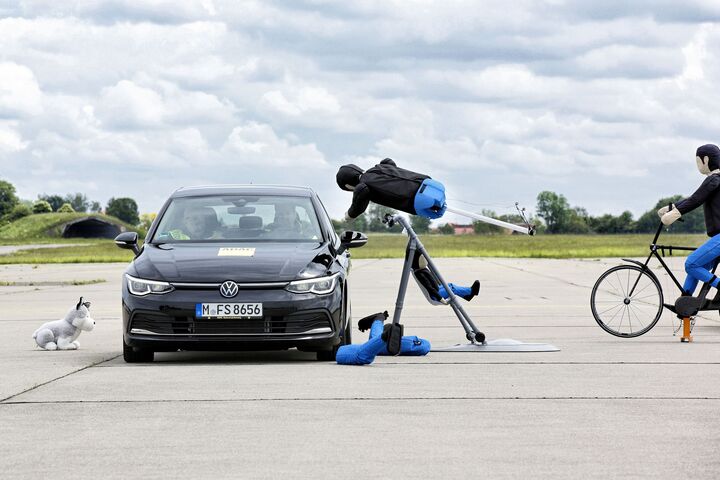 Ein schwarzes Auto am Testgelände. Der Fußgängerdummie fliegt vor dem Auto in die Luft, der Kinderwagen bleibt am Boden. Ein Radfahrerdummie und ein Hundedummie sind neben dem Auto.