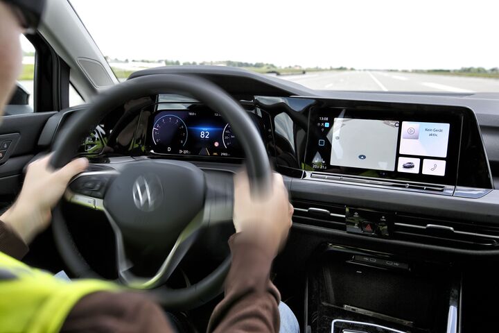 Ein Autofahrer im Cockpit, mit neongelber Weste hält das Lenkrad. Der Tacho zeigt 82 km/h an. Auf der Mittelkonsole befindet sich ein Bildschirm mit Karten- und Fahrzeuginformationen. Durch die Windschutzscheibe ist eine leere, asphaltierte Straße sichtbar.