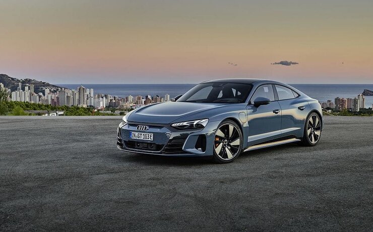 Audi e-Tron GT.jpg Audi e-Tron Sportwagen in blaugrau steht auf einer Straße. Im Hintergrund eine Stadt am Meer und Küste sichtbar.